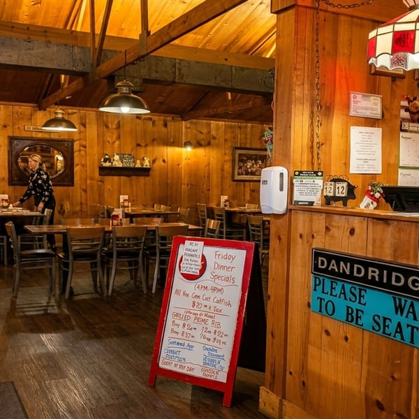 A rustic restaurant interior with wood-paneled walls, tables, chairs, and a hostess stand with a "Please Wait to Be Seated" sign.
