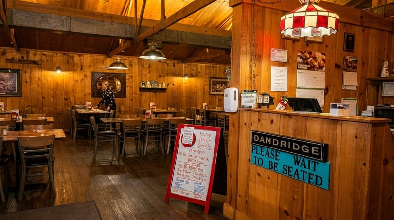 A rustic restaurant interior with wood-paneled walls, tables, chairs, and a hostess stand with a "Please Wait to Be Seated" sign.