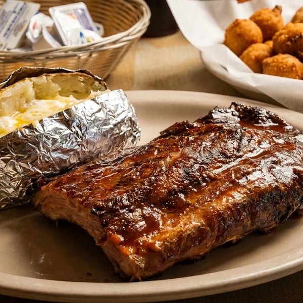 A plate of BBQ ribs with a foil-wrapped baked potato, and a basket of hushpuppies.