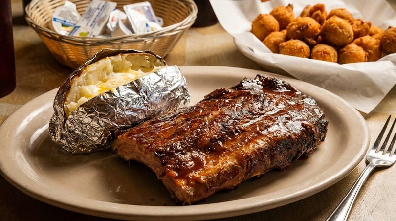 A plate of BBQ ribs with a foil-wrapped baked potato, and a basket of hushpuppies.